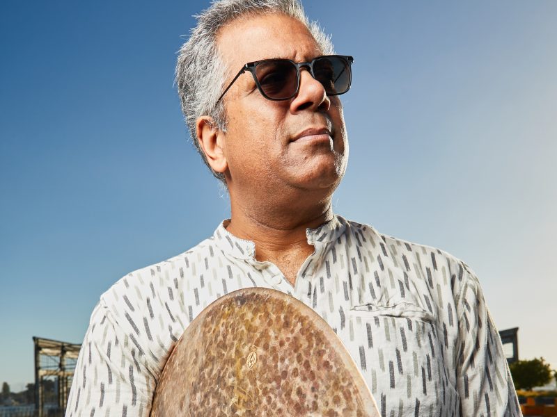 A man with gray hair and sunglasses holds a gong outdoors under a clear blue sky. He is wearing a white shirt with a black pattern.