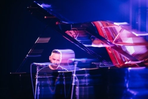 A person plays a grand piano on stage, lit by blue and purple lights, with their reflection visible on the piano's glossy surface.