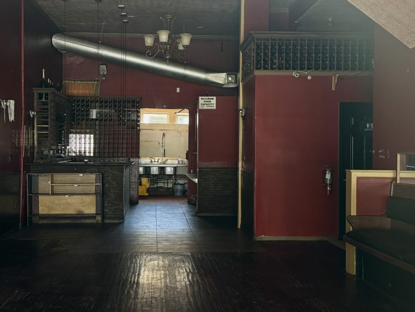 Dimly lit, empty restaurant interior with dark red walls, a wooden bar counter, and an exposed duct on the ceiling.