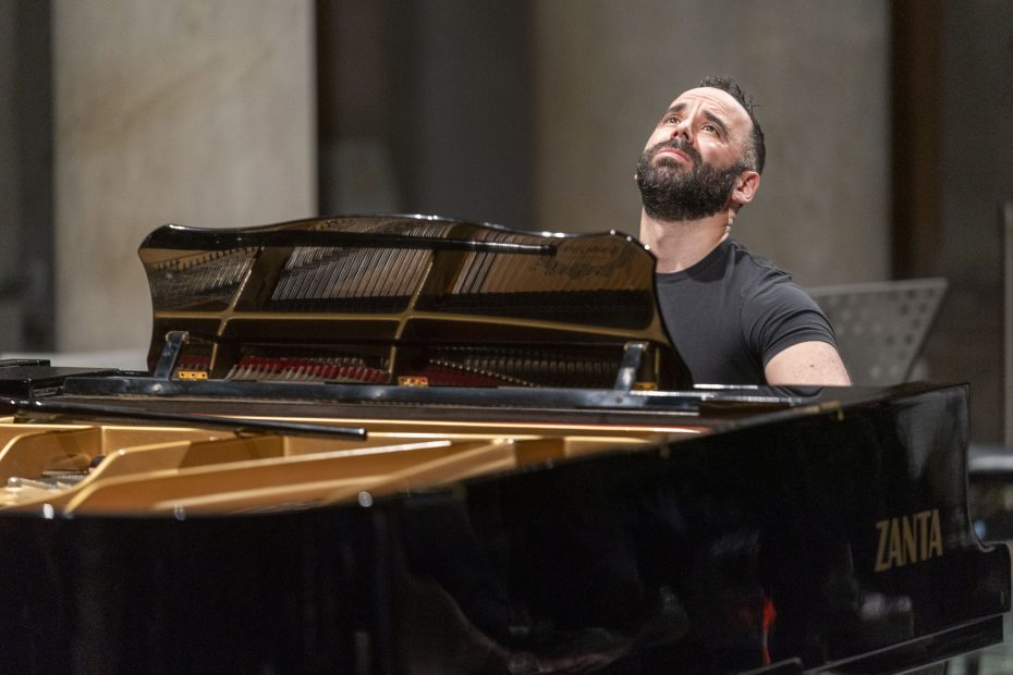 A man with a beard sits at a grand piano, looking upward, with his hands on the keys. The piano lid is open, displaying the strings inside. This scene captures Adam Tendler in his element, immersed in the world of experimental music.