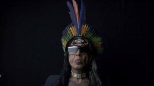 A person wearing a colorful feathered headdress, sunglasses, and a necklace stands against a dark background.