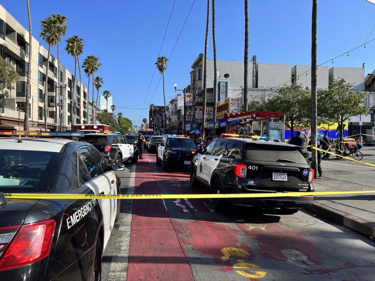 Police and emergency vehicles surround a blocked-off area on a city street lined with buildings and palm trees on a clear day. Police tape restricts access after a shooting.