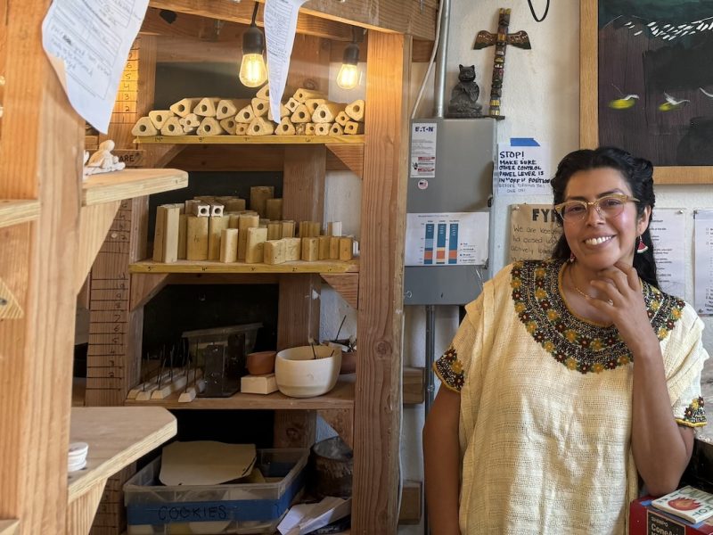 A woman wearing glasses and a colorful embroidered top smiles while standing in a workshop filled with wooden shelves, tools, and various supplies, amid the rustic charm of her ag workshop.