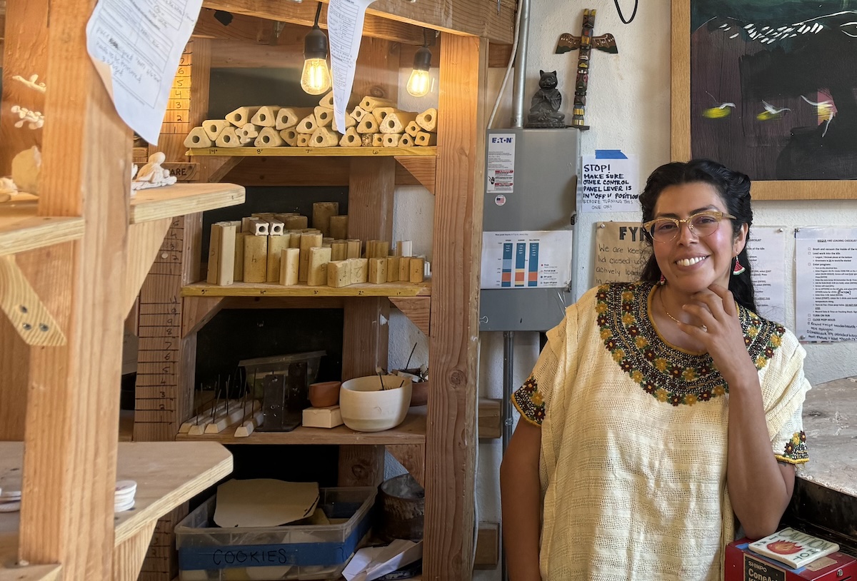 A woman wearing glasses and a colorful embroidered top smiles while standing in a workshop filled with wooden shelves, tools, and various supplies, amid the rustic charm of her ag workshop.