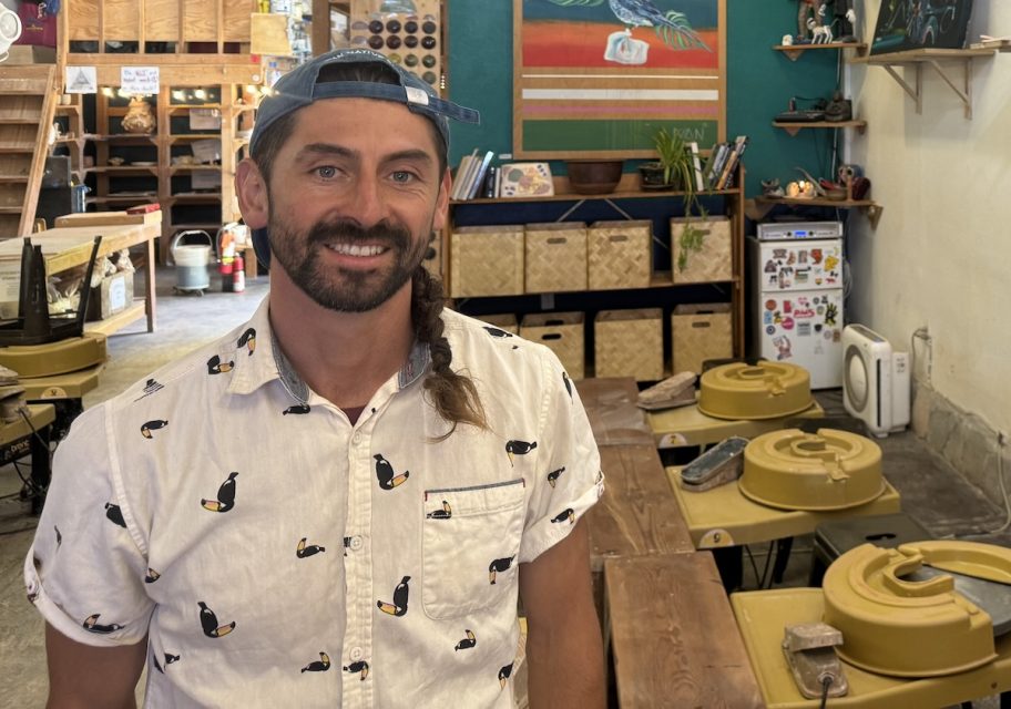 A man sporting a toucan-print shirt and a backwards cap stands inside a pottery studio, with pottery wheels and various equipment, reminiscent of an artistic form of artillery, in the background.