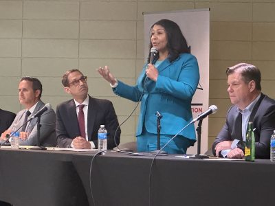 A woman in a blue suit speaks into a microphone on a panel with four other individuals seated at a table with microphones and water bottles.