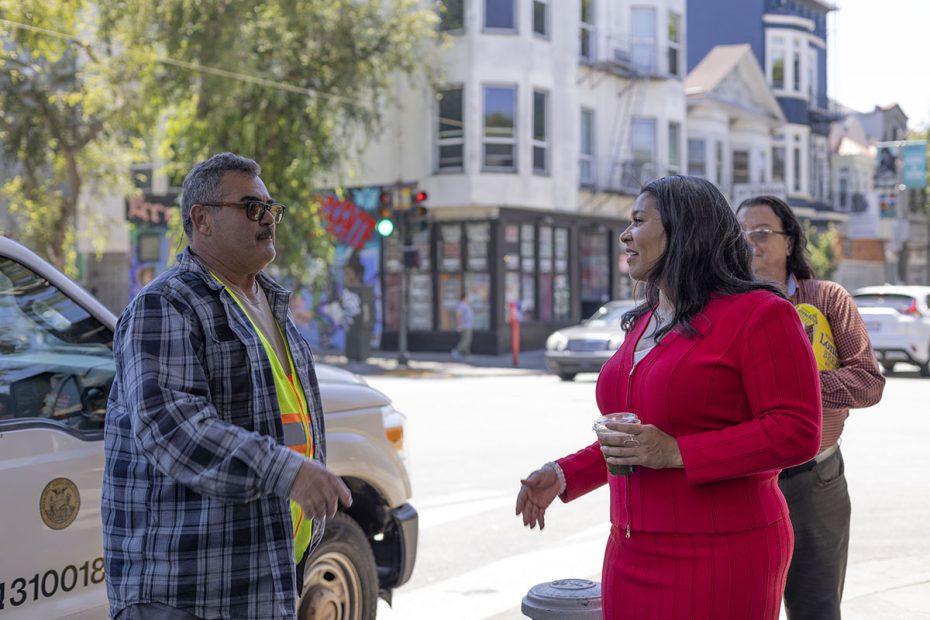 Three people stand on a city street corner near a parked van with a municipal logo. One person in a red outfit holds a drink, while another in a plaid shirt and reflective vest talks and gestures.