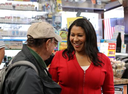 A woman in a red jacket smiles while talking to an older man in a baseball cap inside a store with an American flag in the background.