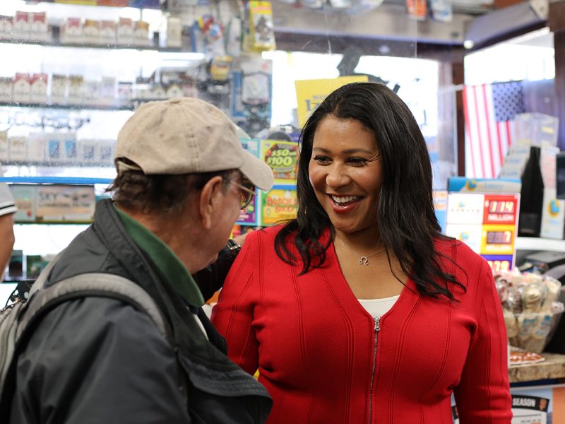 A woman in a red jacket smiles while talking to an older man in a baseball cap inside a store with an American flag in the background.