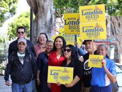 A diverse group of people poses outdoors, holding yellow signs supporting Mayor London Breed, who stands in the center wearing a red dress.