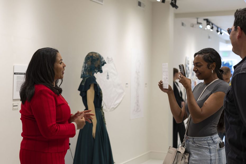 Three people stand in an art gallery. One person in a red outfit talks while another person in a grey outfit takes a photo. Art pieces, including a mannequin in a dress, are displayed on the walls.