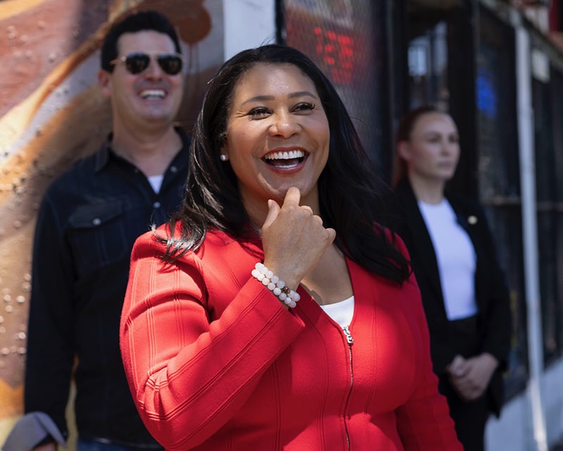 Mayor London Breed in a red jacket stands smiling with her hand near her face, accompanied by two people in the background.