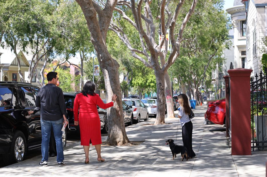 Three people and a small dog are on a sidewalk lined with trees and parked cars. A woman in a red dress waves at another woman walking a dog. A man stands nearby holding a face mask.