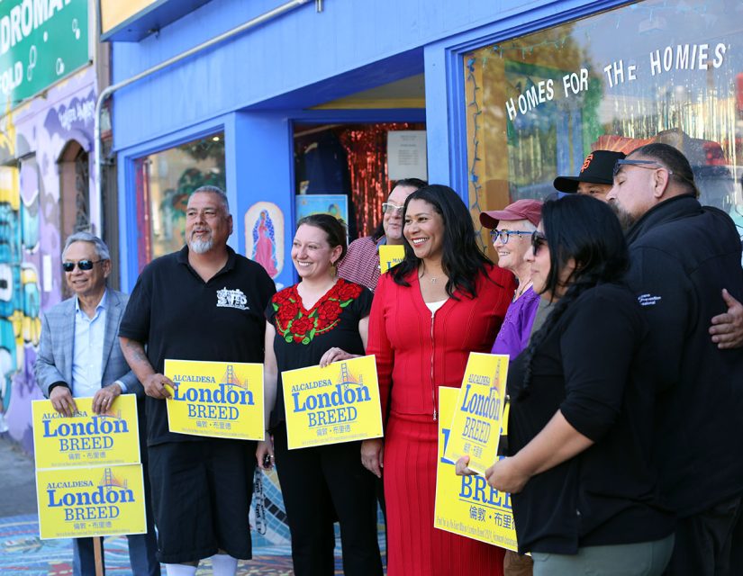 Group of people, including a woman in a red dress, standing outside a colorful building holding yellow signs that read "London Breed" and "Alcaldesa.