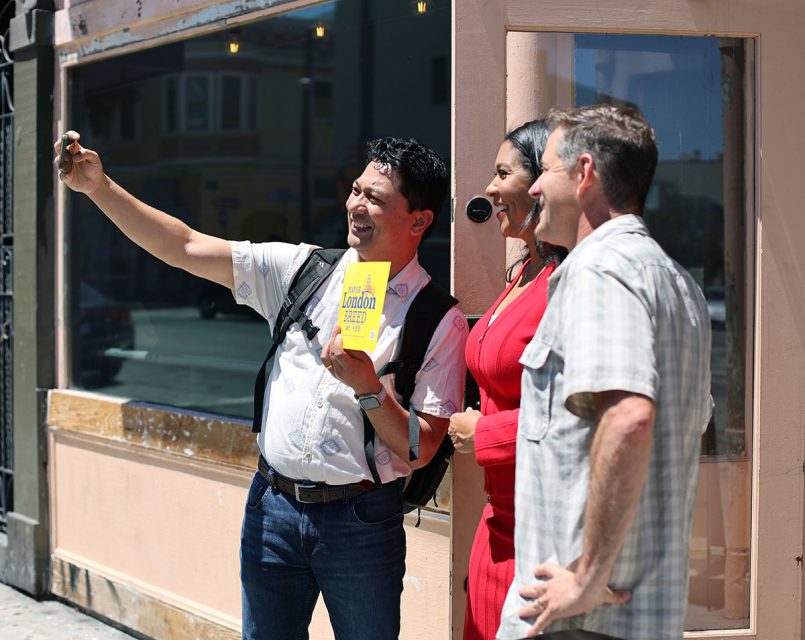 Three people stand outside a building, posing for a selfie. The person on the left holds a London travel guidebook.