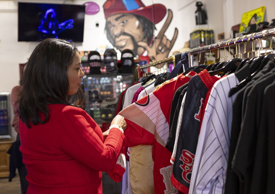 Person in a red jacket browsing through jerseys in a clothing store adorned with a mural and sports memorabilia.