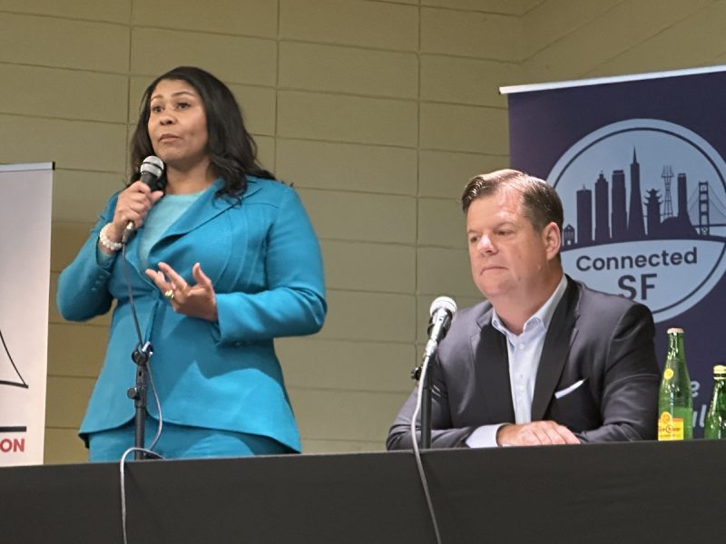 A woman in a blue blazer speaks into a microphone while standing, next to a seated man in a dark suit at a conference table. A "Connected SF" banner is in the background.