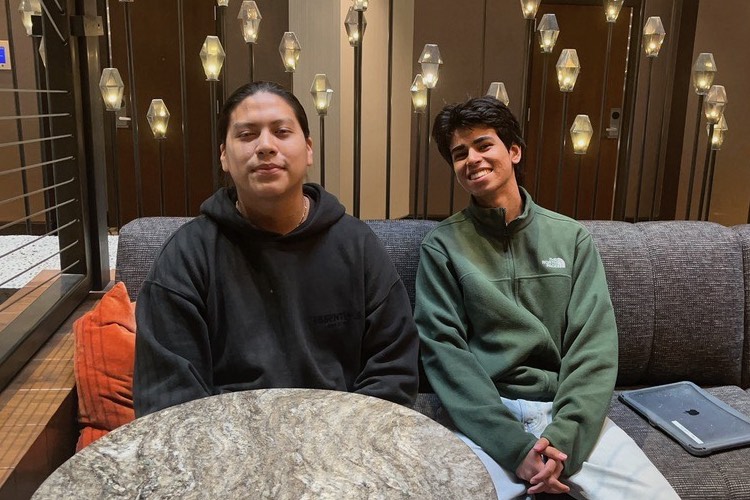 Two individuals sit on a couch in a modern setting with decorative lighting in the background; one wears a black hoodie and the other a green jacket. A round marble table is in front of them.