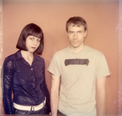 Two individuals stand side by side against a plain background. The person on the left has dark shoulder-length hair and a dark shirt. The person on the right has short hair and is wearing a light-colored T-shirt.