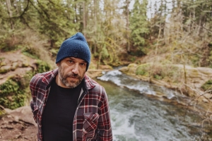 A person in a plaid shirt and blue beanie stands near a river with flowing water, surrounded by a forest.