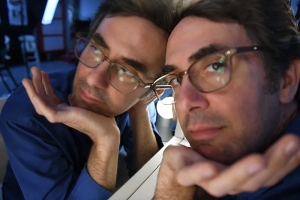 A person wearing glasses rests their hands on their cheeks and looks into a mirror, creating a reflection of their face.