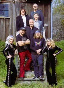 Seven people, three seated on outdoor steps and four standing, pose in front of a wooden structure.