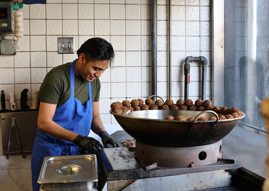 A person in a blue apron and green shirt prepares food beside a large frying pan filled with spherical food items in a tiled kitchen.