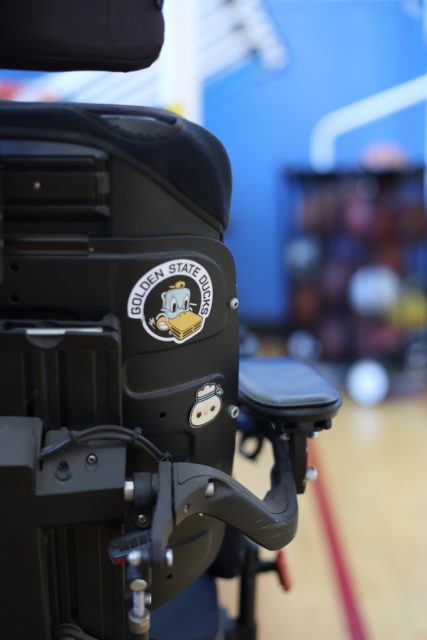 Close-up of a wheelchair adorned with a sticker featuring a cartoon duck and text reading "Golden State Ducks," positioned in an indoor setting with blurred gymnasium equipment in the background.