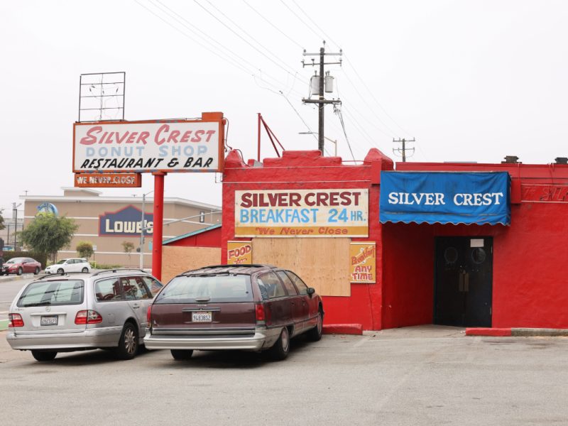 A red building houses the Silver Crest Donut Shop, Restaurant, and Bar. Two cars are parked in front. Signs advertise 24-hour breakfast and an always open status.