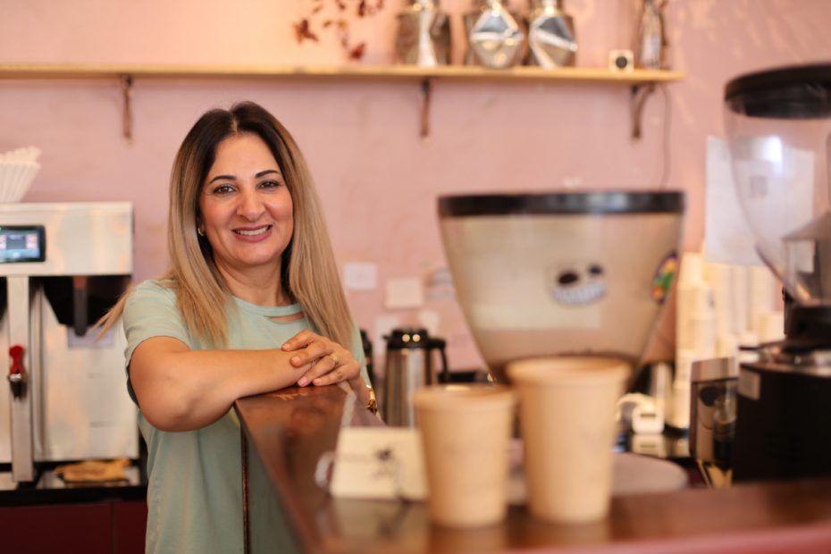 A woman in a green top stands behind a counter in a coffee shop, smiling at the camera. In front of her, there are three brown paper cups. Coffee-making equipment is visible in the background.