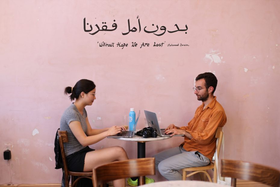 Two people sit across from each other at a table in a pink-walled cafe, working on laptops. A camera is on the table. A quote in Arabic and English is on the wall.