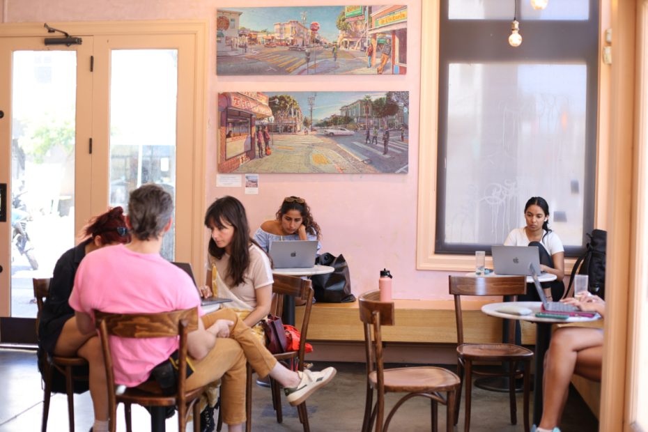 People sitting at tables, working on laptops, and conversing in a cozy cafe with pastel walls and artwork hanging on the walls.