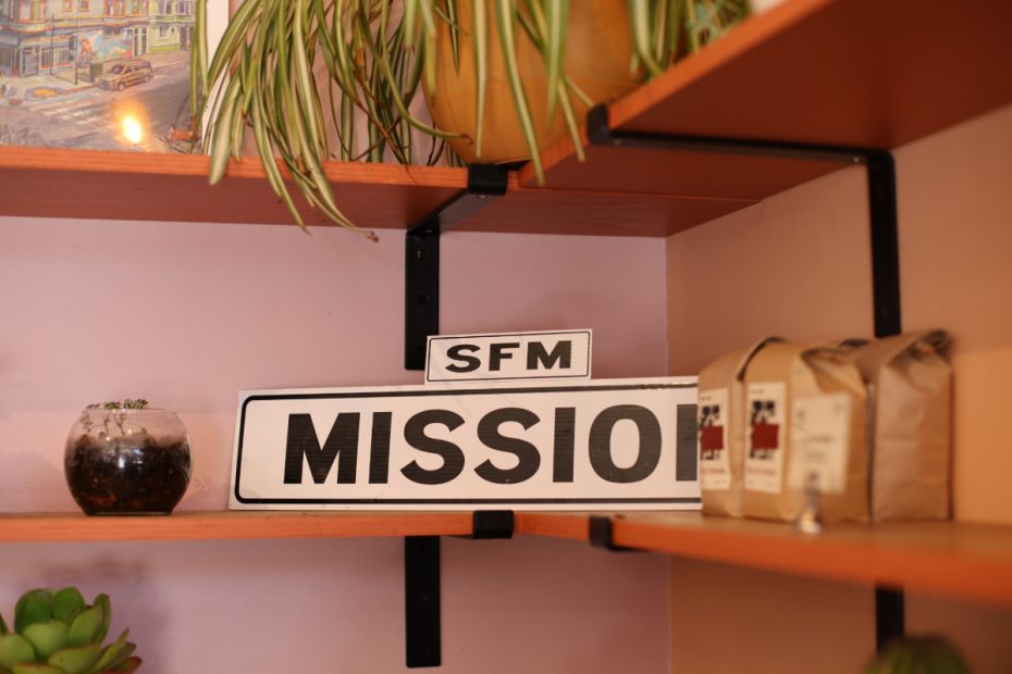 A shelf with plants, a painting, and a "SFM Mission" street sign. Several brown paper coffee bags are arranged next to the sign.