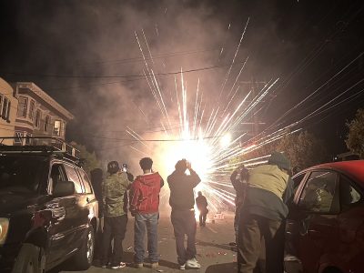 A group of people stands on a street at night, watching July 4 fireworks light up the distance.