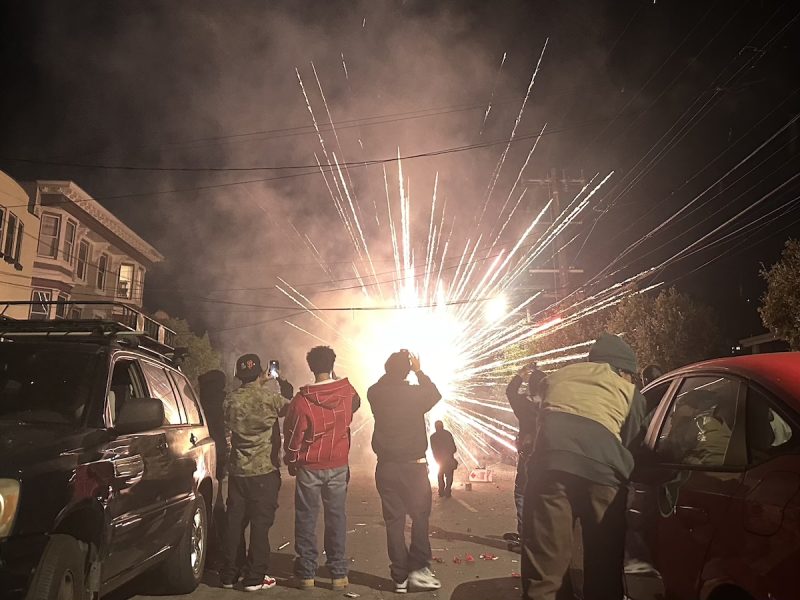 A group of people stands on a street at night, watching July 4 fireworks light up the distance.