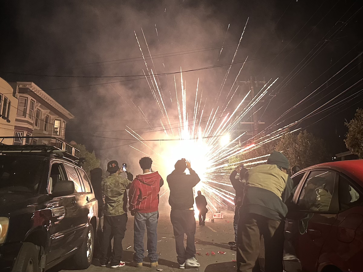 A group of people stands on a street at night, watching July 4 fireworks light up the distance.