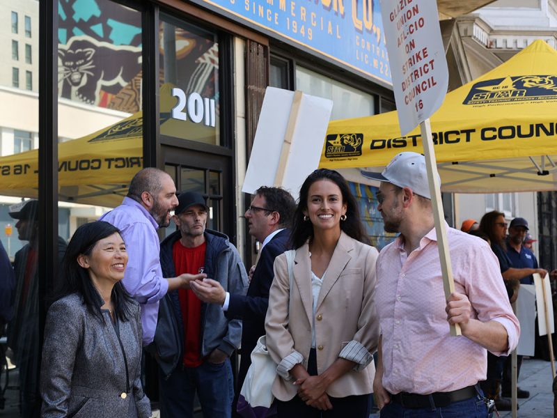 People stand and talk outside a commercial building with yellow tents and signs. One person holds a protest sign.
