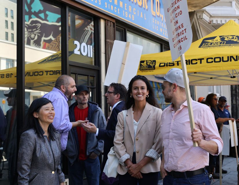 People stand and talk outside a commercial building with yellow tents and signs. One person holds a protest sign.