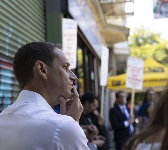 A man in a white shirt appears deep in thought while several people in the background hold protest signs.