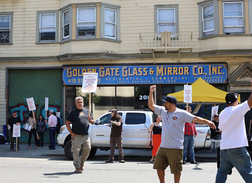 A group of people with picket signs are gathered outside Golden Gate Glass & Mirror Co., Inc. building. A man in the foreground gestures while holding a sign.