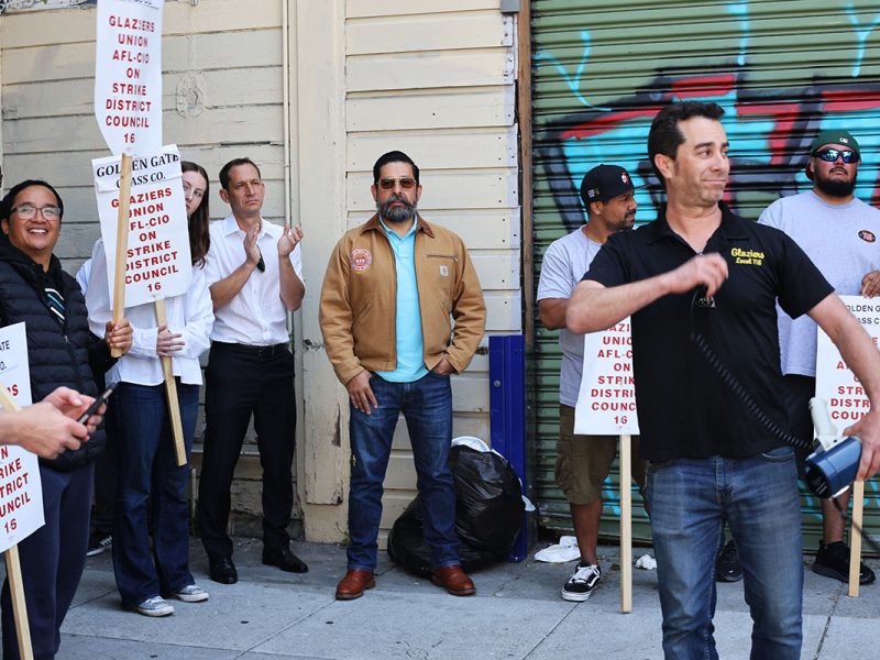 A group of people, some holding signs reading "Union on Strike District Council 16," stand together in front of a building with a closed, graffiti-covered security shutter.