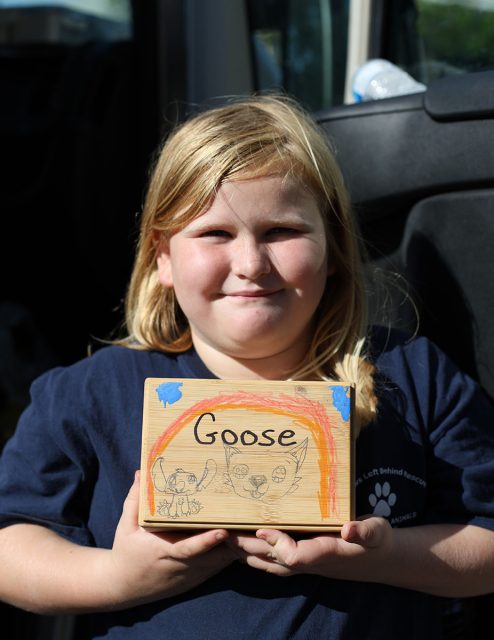 A child holds a rectangular wooden box labeled "Goose" with an illustrated cat face and other small doodles. The child is standing in front of an open vehicle door.