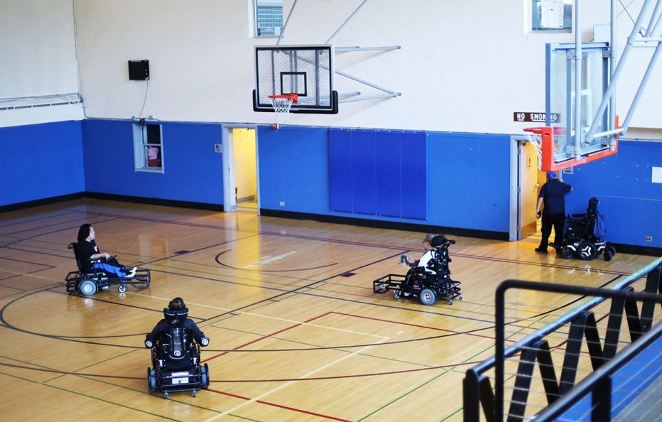 Four individuals in electric wheelchairs play a game of wheelchair basketball in a gymnasium with blue walls and a wooden floor.