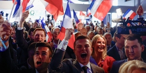 A group of enthusiastic people cheer and wave flags in a crowded indoor event.