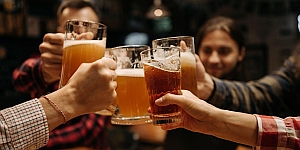 Four people clink glasses filled with beer in a toast, with a dimly-lit background suggesting a casual setting like a bar or pub.