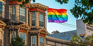 A rainbow Pride flag is displayed on the exterior of a multi-story building in an urban setting with trees partially framing the image.