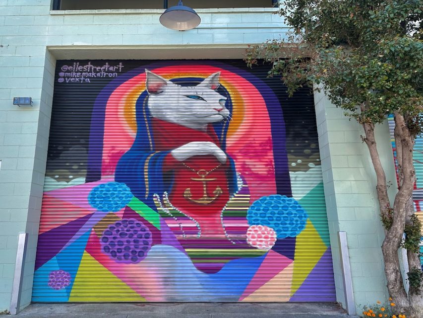 A mural on a building depicts a cat wearing a robe, holding a heart with an anchor symbol. The background features colorful geometric shapes and patterns.