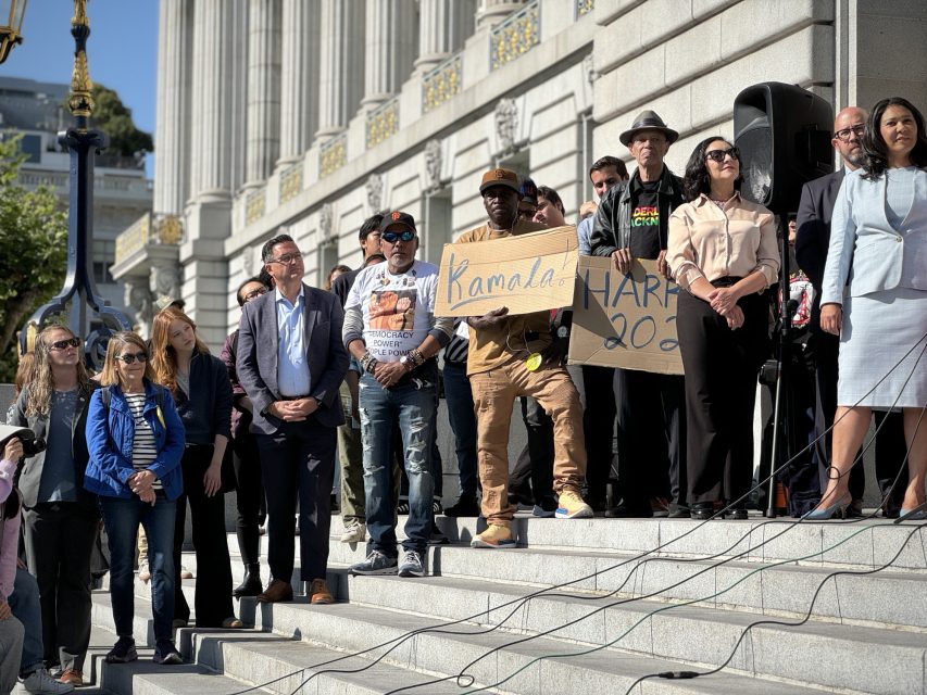 A group of people stands on the steps of a large building for a public event. One person is holding a cardboard sign that reads "Kamala Harris 2024".