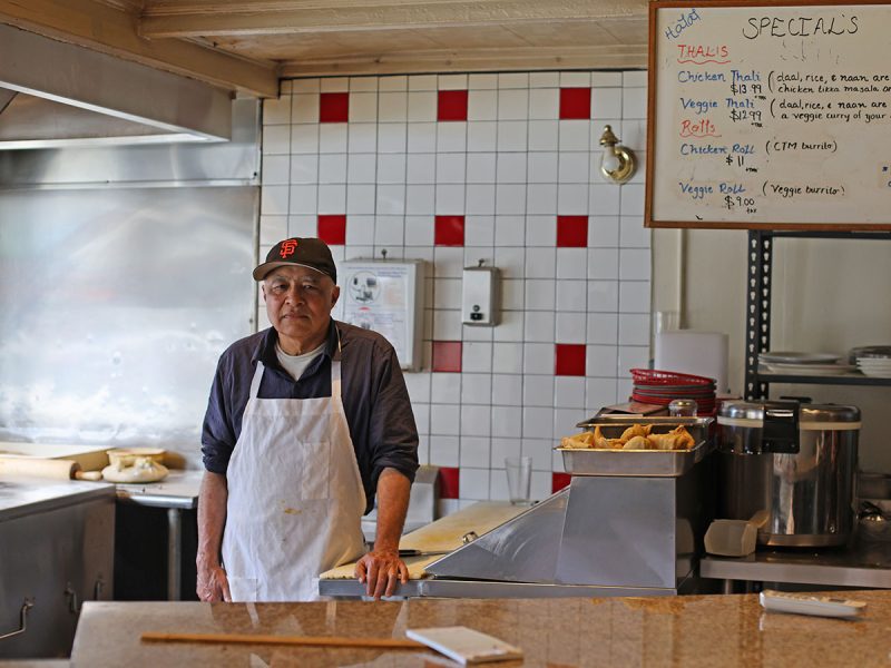 Man wearing a white apron and cap stands behind a counter in a kitchen with red and white tiles. A specials menu is visible to the right, and kitchen equipment and prepared food can be seen.