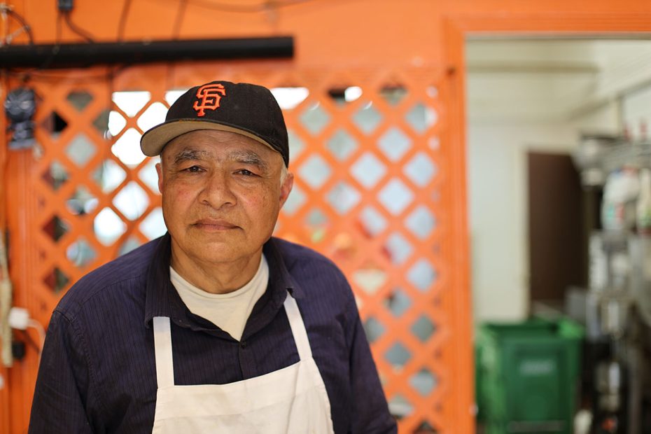 A man wearing a San Francisco Giants cap and an apron stands in front of an orange lattice background inside a building.
